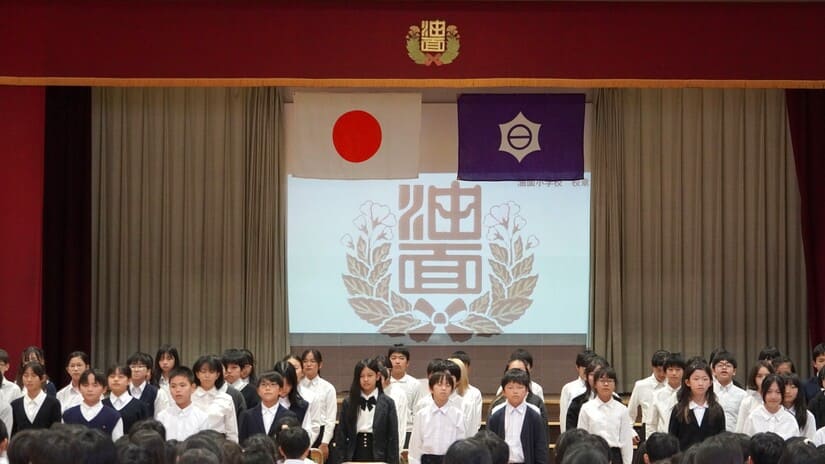 真剣な表情で開校100年のあゆみを発表する油面小6年生の皆さん。ステージ上のスクリーンには油面小の校章が映し出されています。