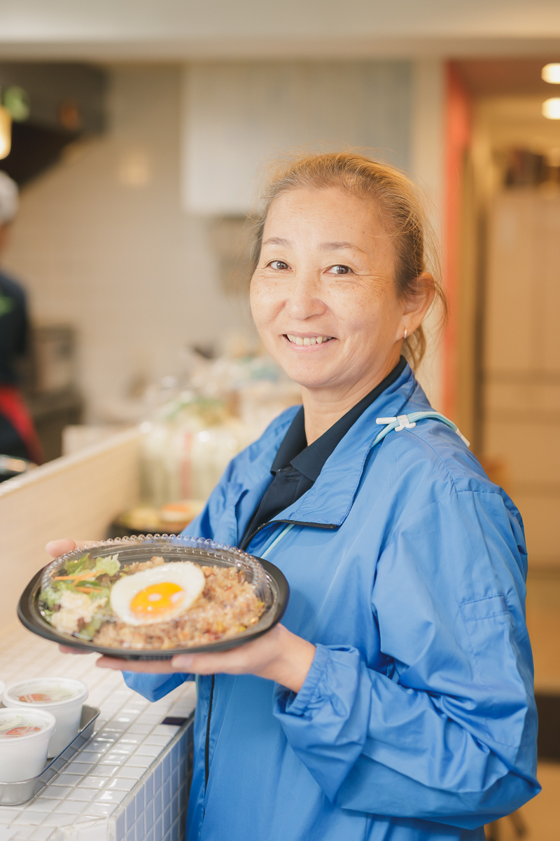 代表者の芹田明美さんの写真。ナシゴレン弁当を両手で持って微笑んでいます。