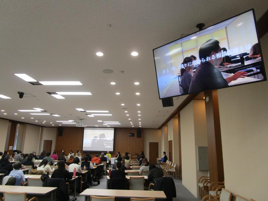 当日の様子。大会議室に長机が並んでいる。参加者は前方の発表者やスクリーンを見ている。